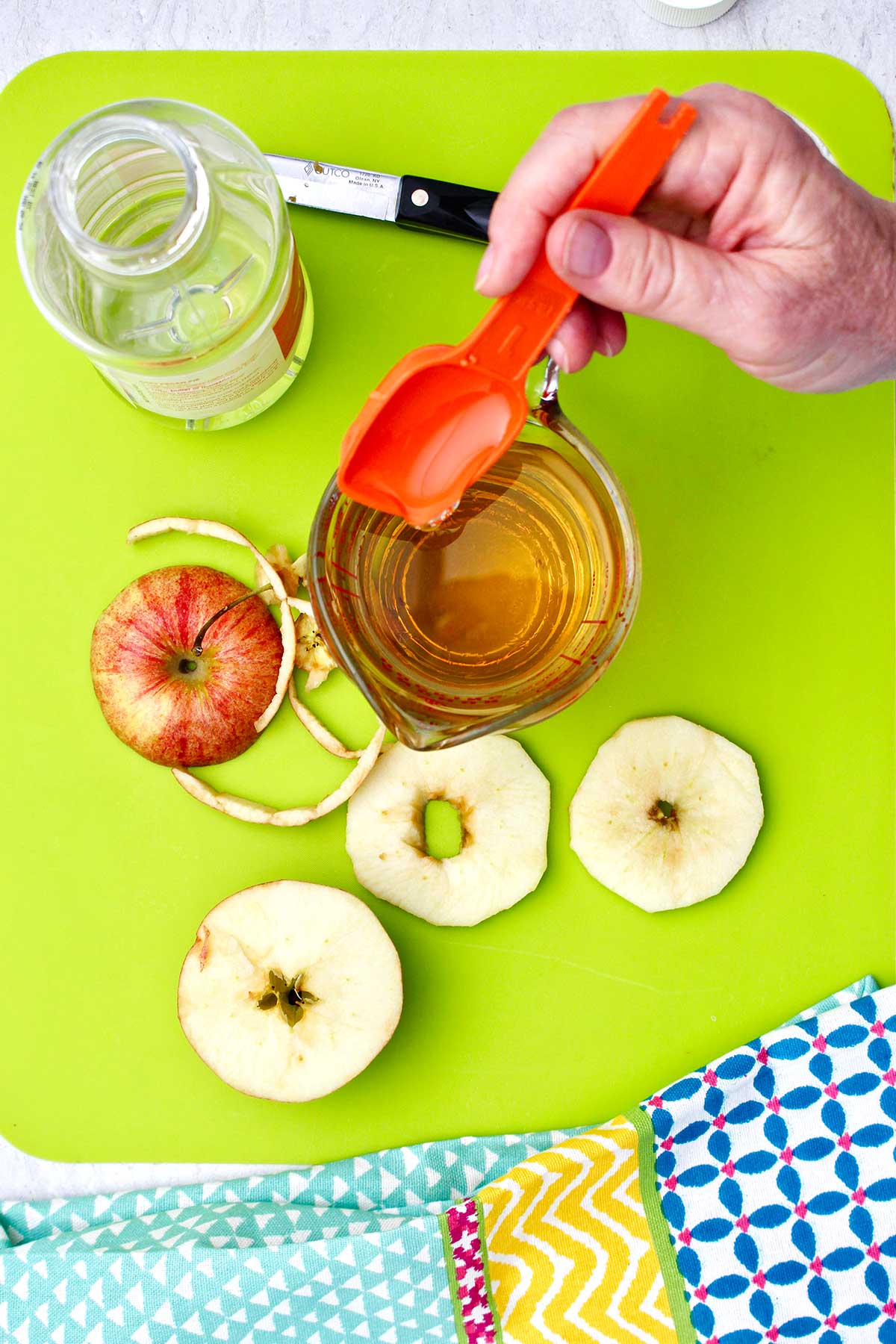 Person pouring honey into a glass measuring cup with slices of apples near by on a bright green cutting board.