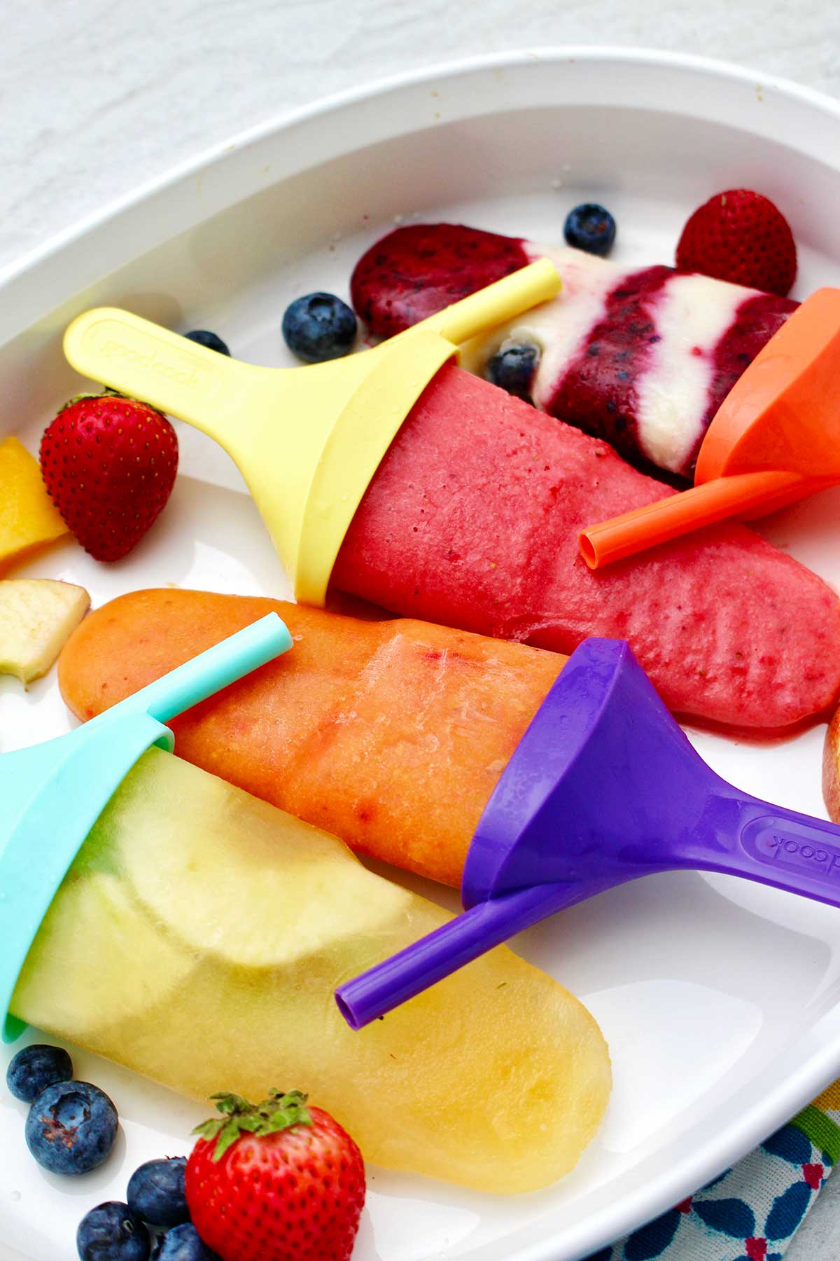 Four different flavored popsicles on colorful popsicle holders resting on a platter of fruit.