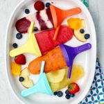 Four different flavored popsicles on colorful popsicle handles resting on a platter of fruit.