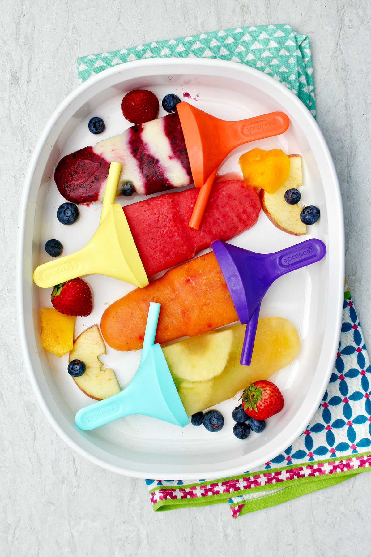 Four different flavored popsicles on colorful popsicle holders resting on a platter of fruit.