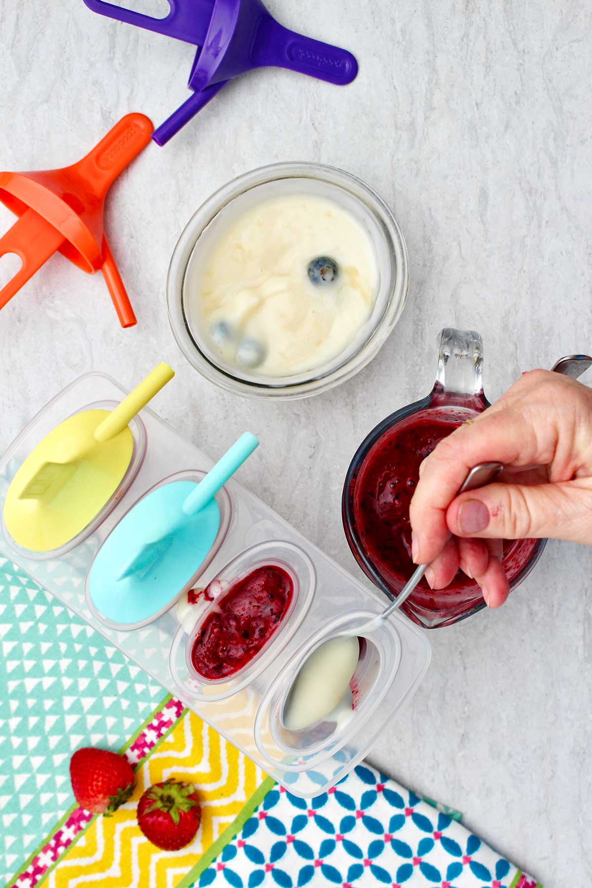 Person spooning blueberry yogurt into popsicle mold with other supplies near by.