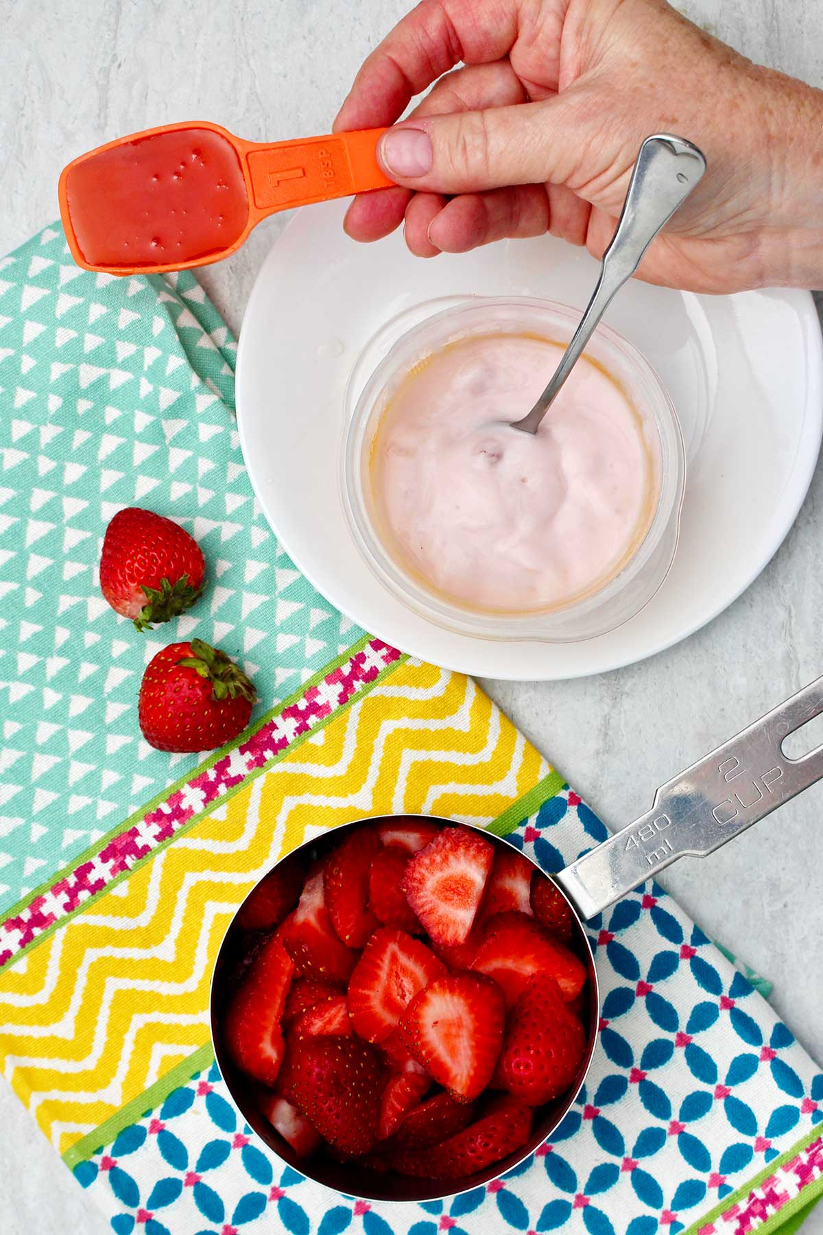 Person holding an orange plastic tablespoon of honey near a cup of strawberries and cup of yogurt.