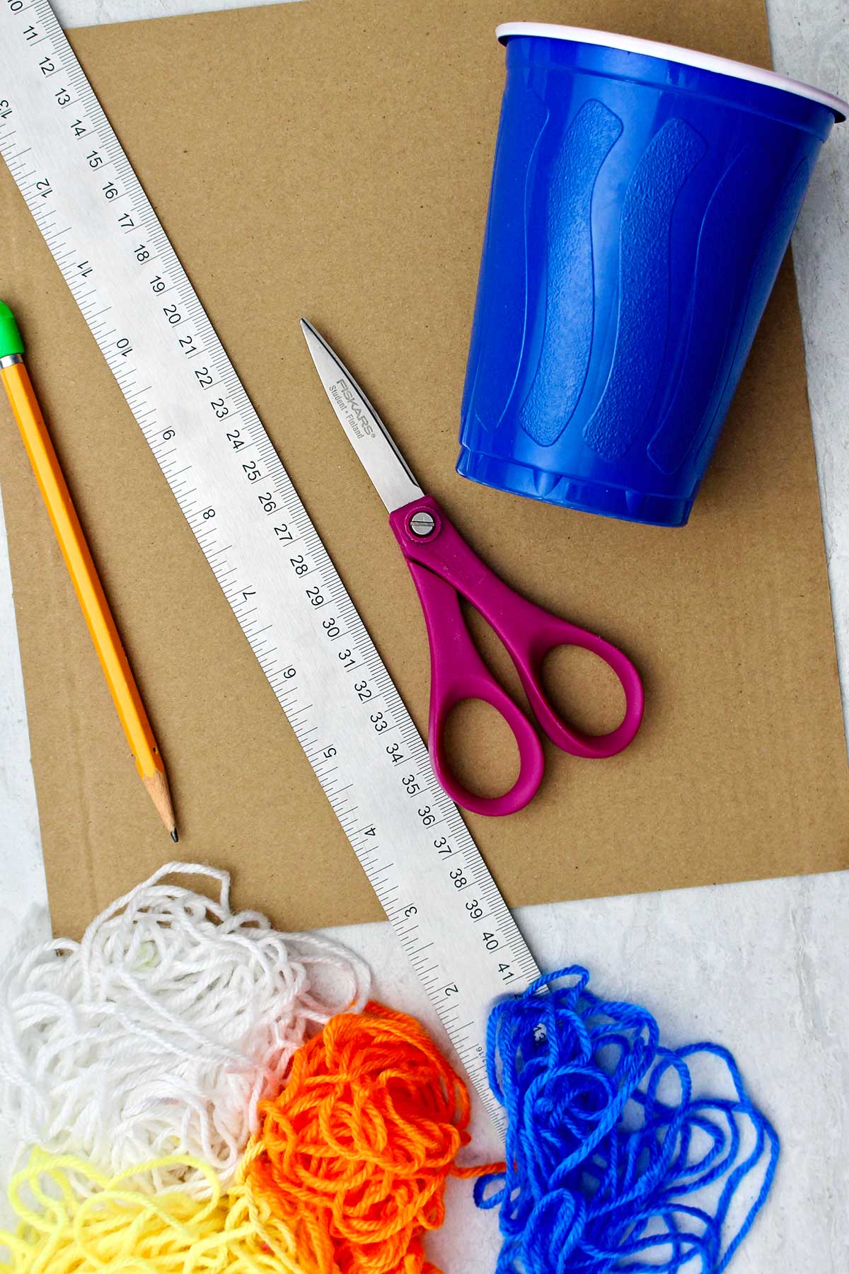 Supplies for making round friendship bracelets. Metal ruler, cup, scissors, cardboard, pencil, colorful bundles of yarn.