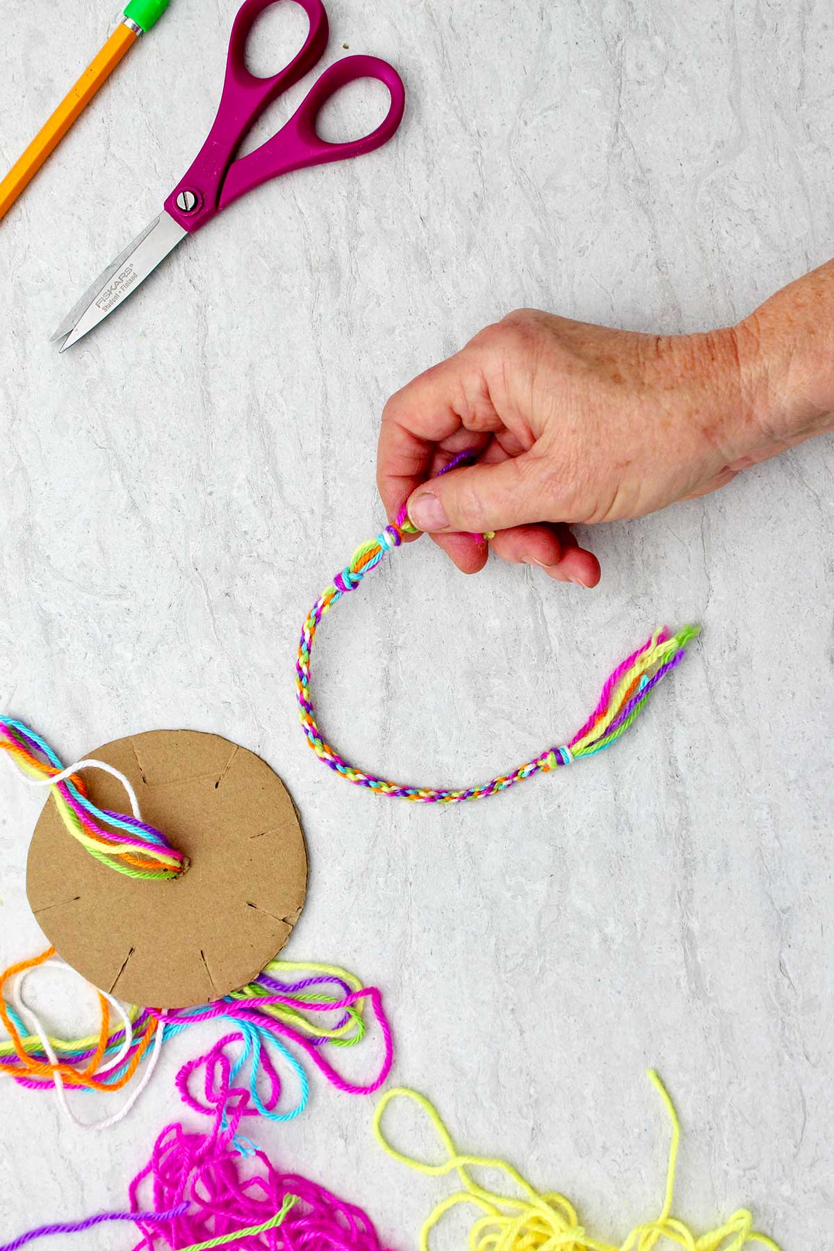 Person holding the end of a completed round friendship bracelet, scissors, pencil, cardboard loom and colorful yarn.