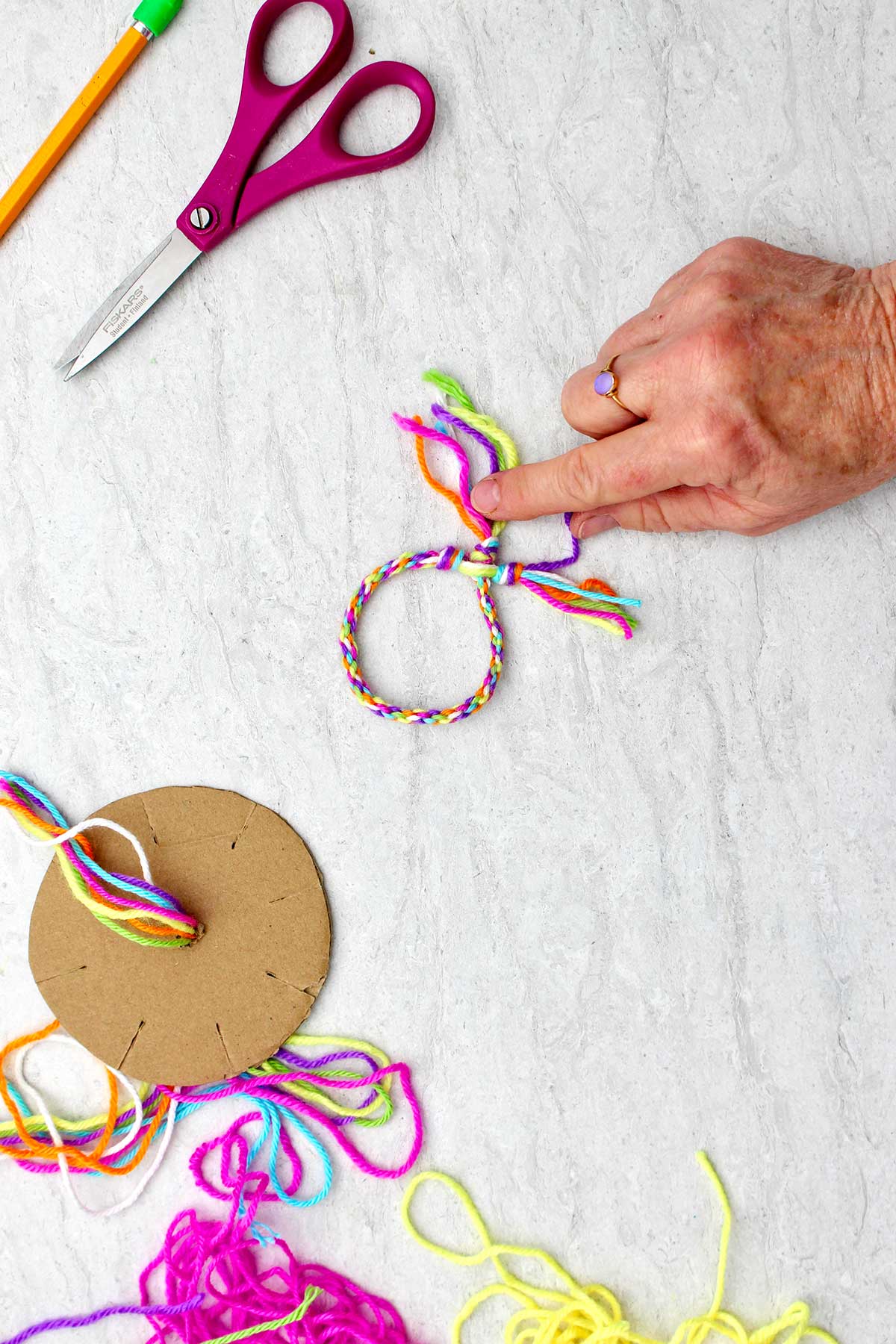 Completed round friendship bracelet, scissors, pencil, cardboard loom and colorful yarn.