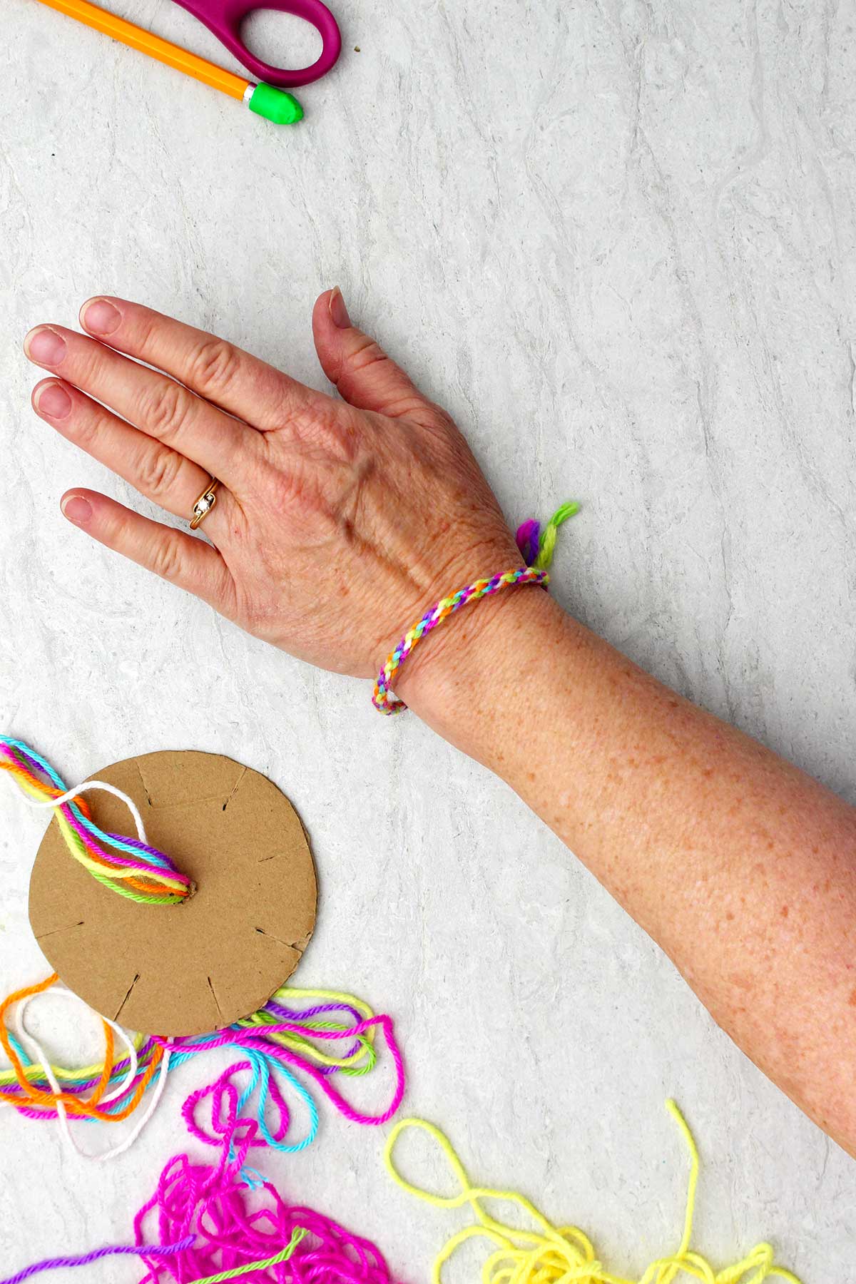 Completed round friendship bracelet on a woman's wrist with a cardboard loom and yarn near by.