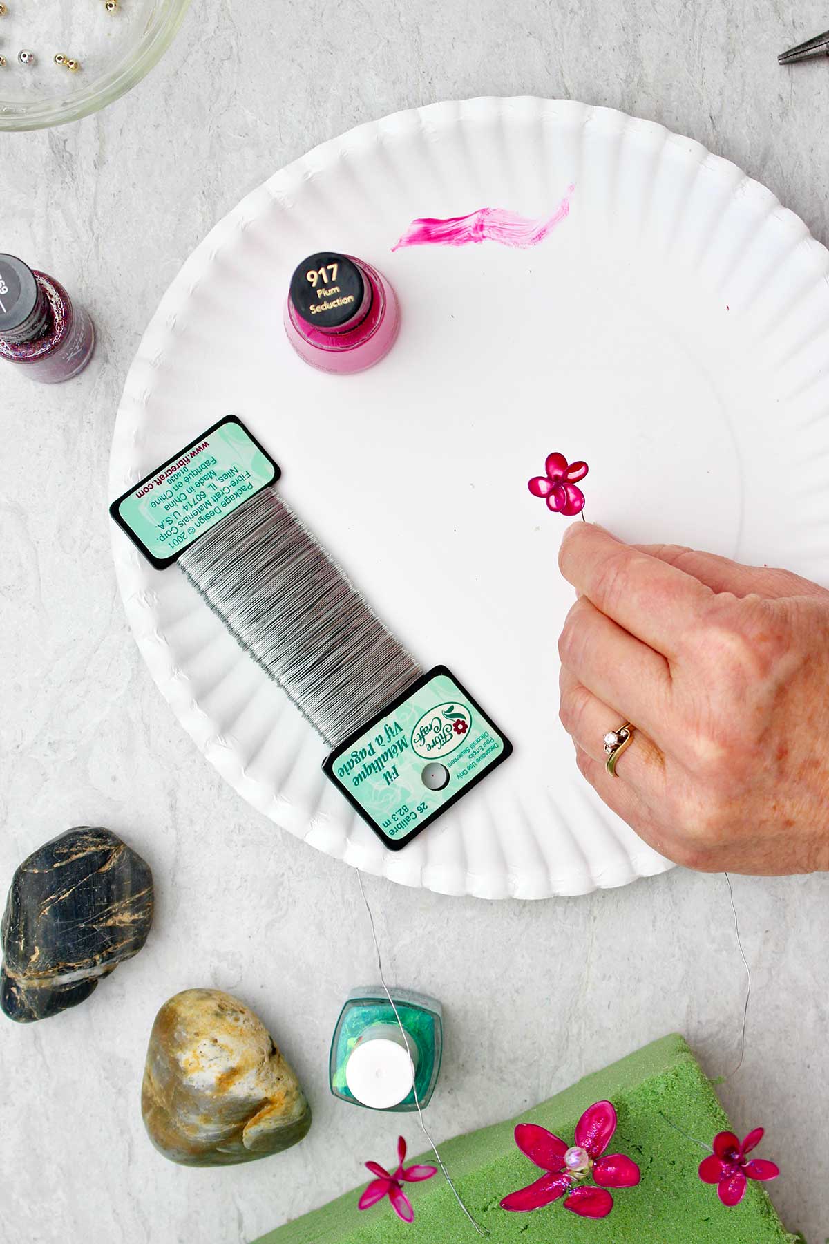 Person holding pink nail polish and wire flower over a white paper plate with other supplies near by.