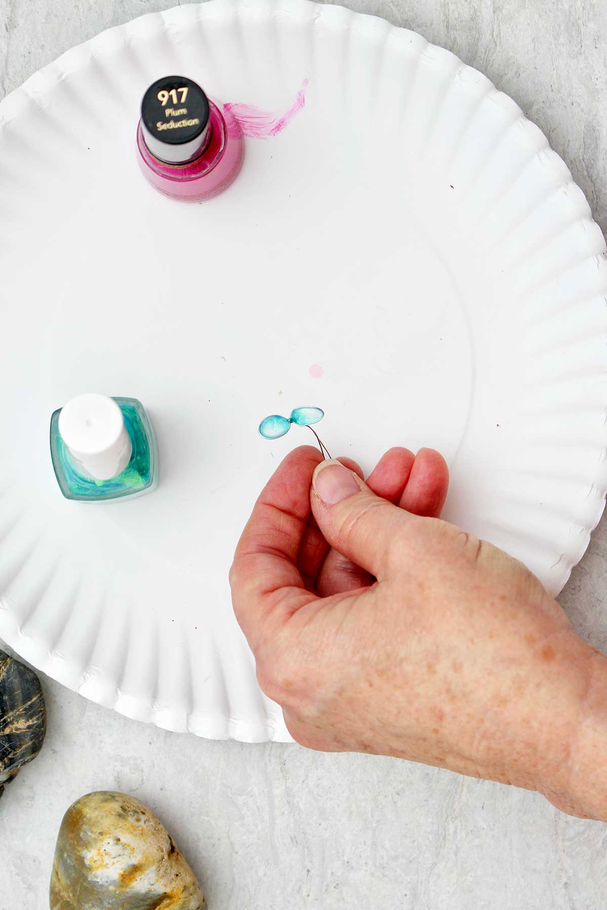 Person holding blue nail polish and wire flower over a white paper plate.