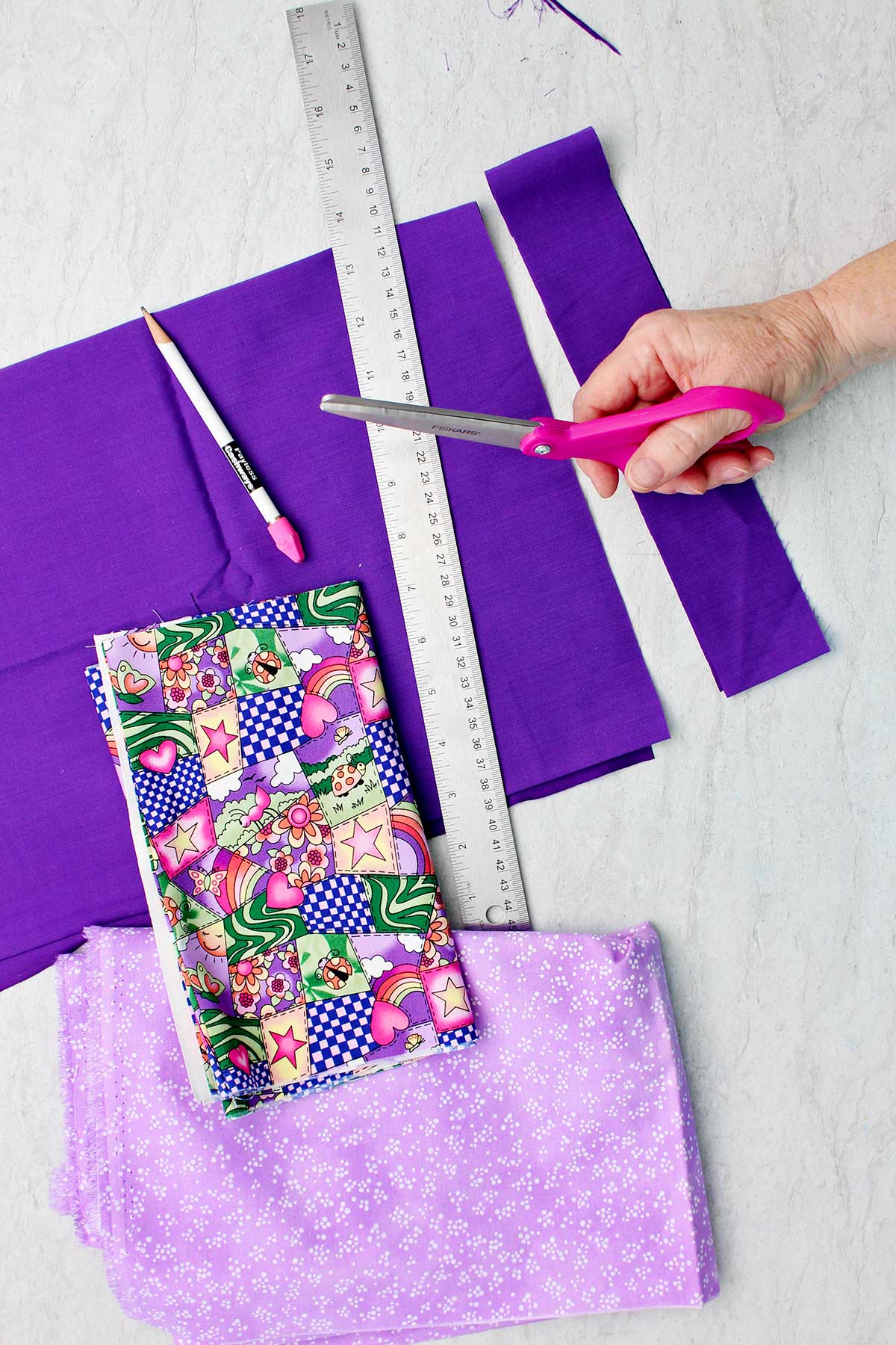 Person holding pink scissors, ruler, pins, and various purple fabrics.