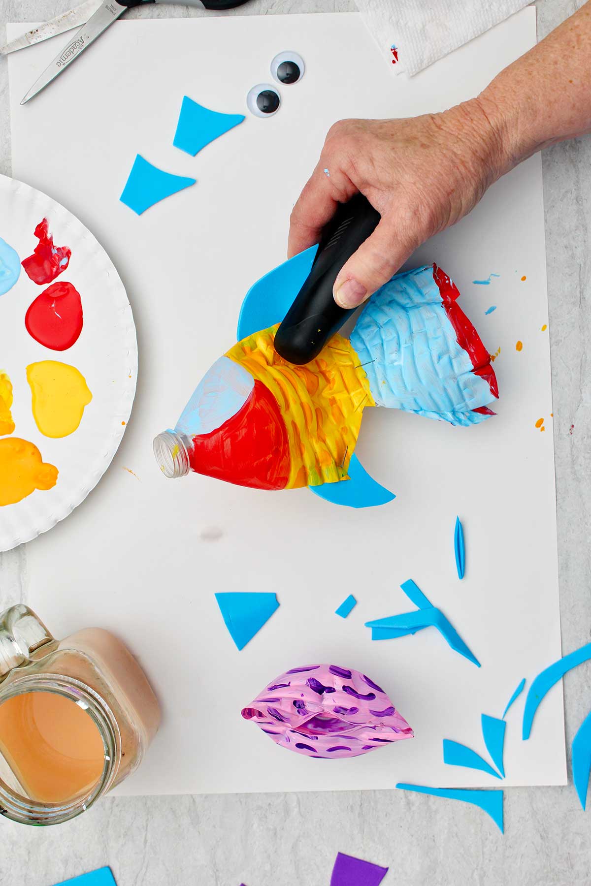 Person stapling blue foam fin to painted water bottle fish.