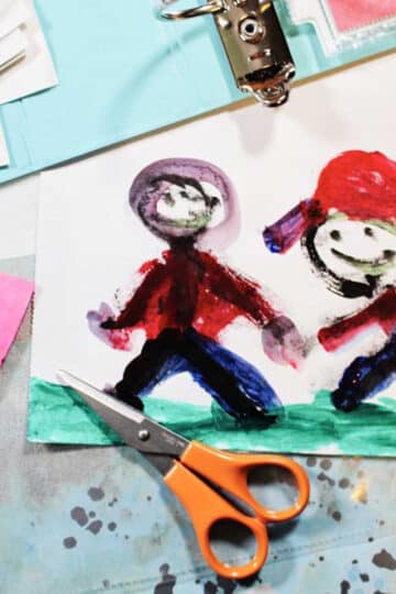 A child's painting of two people holding hands, with scissors, a binder and scrapbook paper nearby.