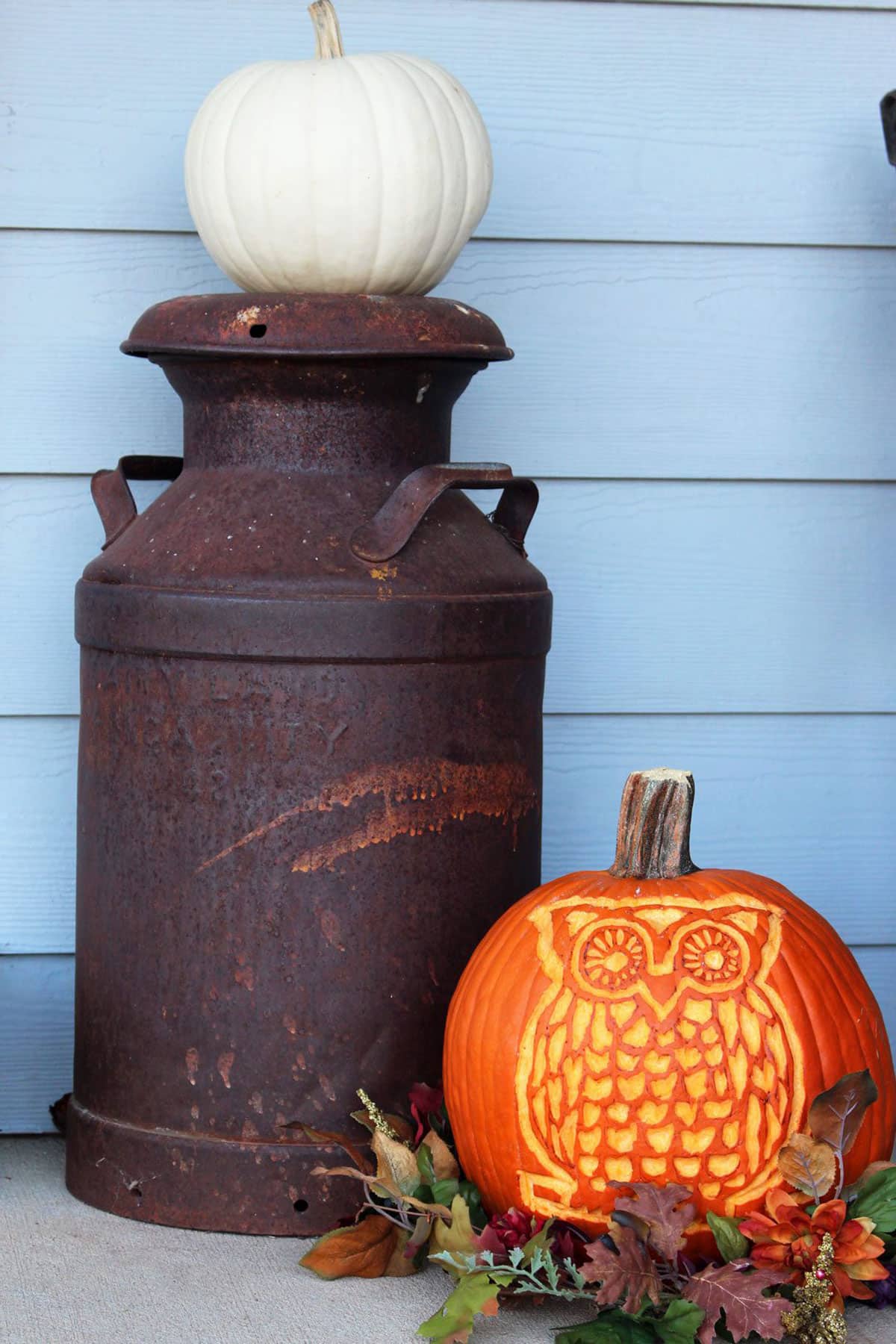 An owl carved pumpkin sitting with some fall leaves and other fall decorations on a front porch.