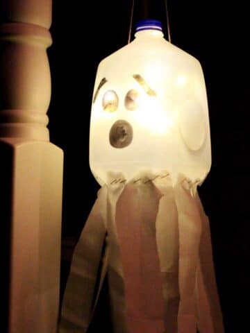 A recycled milk jug with a face drawn on it to create a glowing ghost luminary hanging on a porch at night.