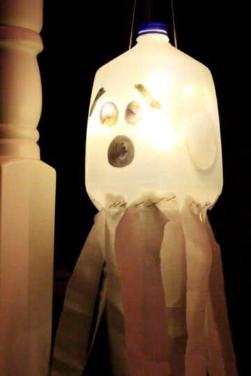 A recycled milk jug with a face drawn on it to create a glowing ghost luminary hanging on a porch at night.