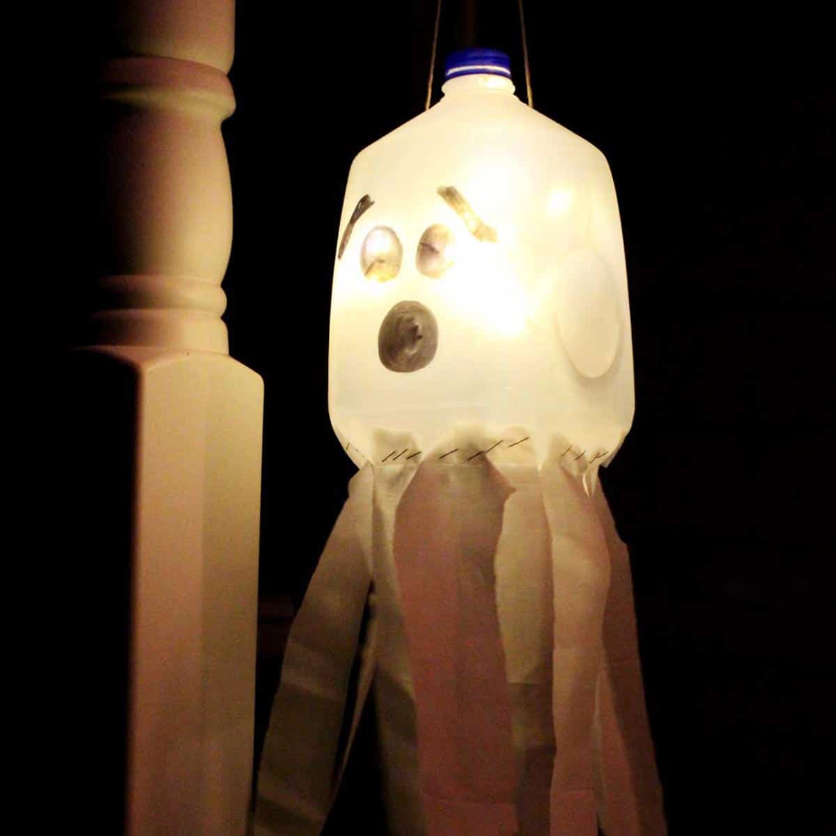 A recycled milk jug with a face drawn on it to create a glowing ghost luminary hanging on a porch at night.