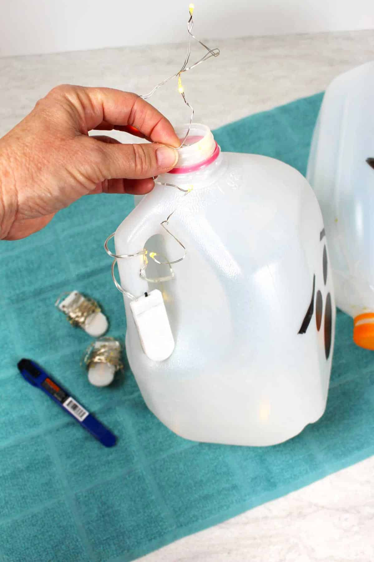 A string of lights being placed inside a recycled milk jug with a ghost face drawn on the outside.