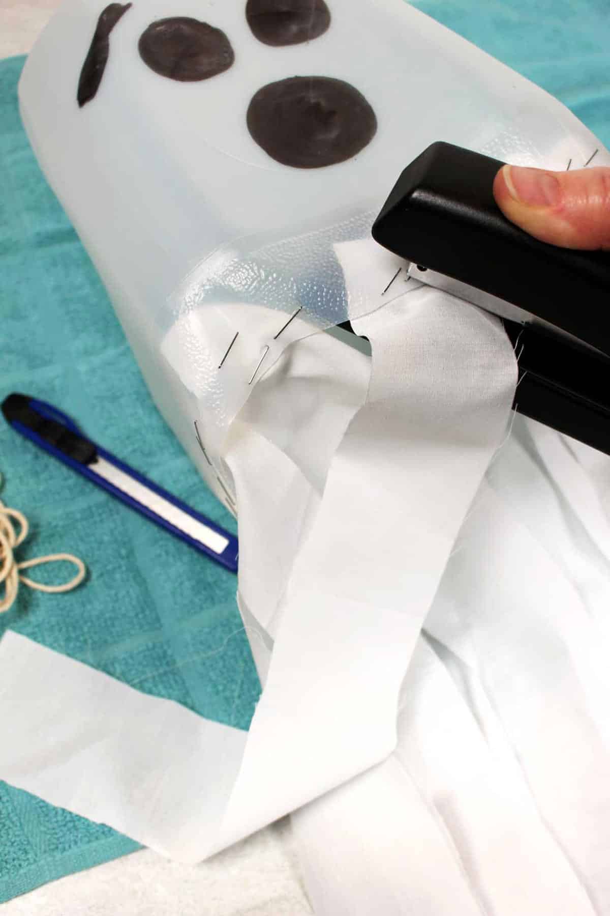 A stapler attaching strips of white cloth to the bottom of a milk jug ghost.