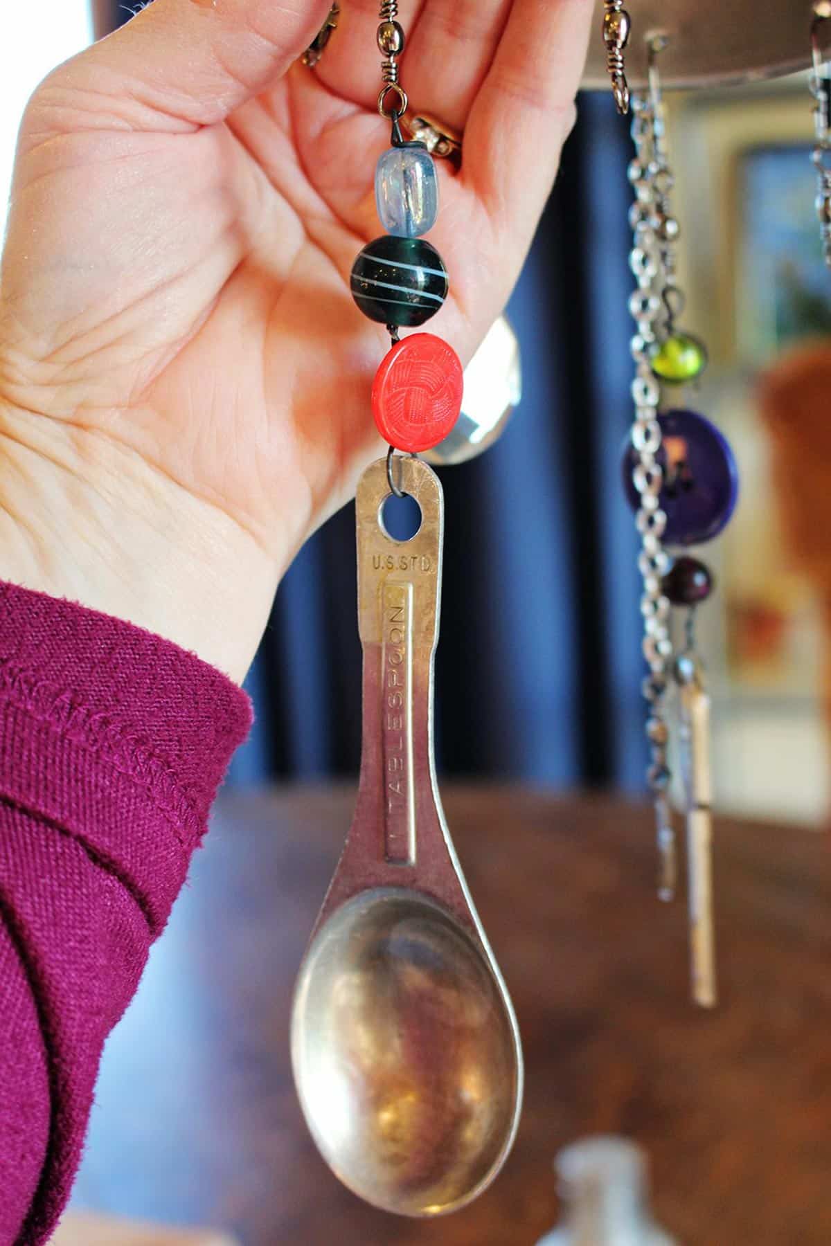 A hand holding a vintage measuring spoon with a button and beads strung on a wire.