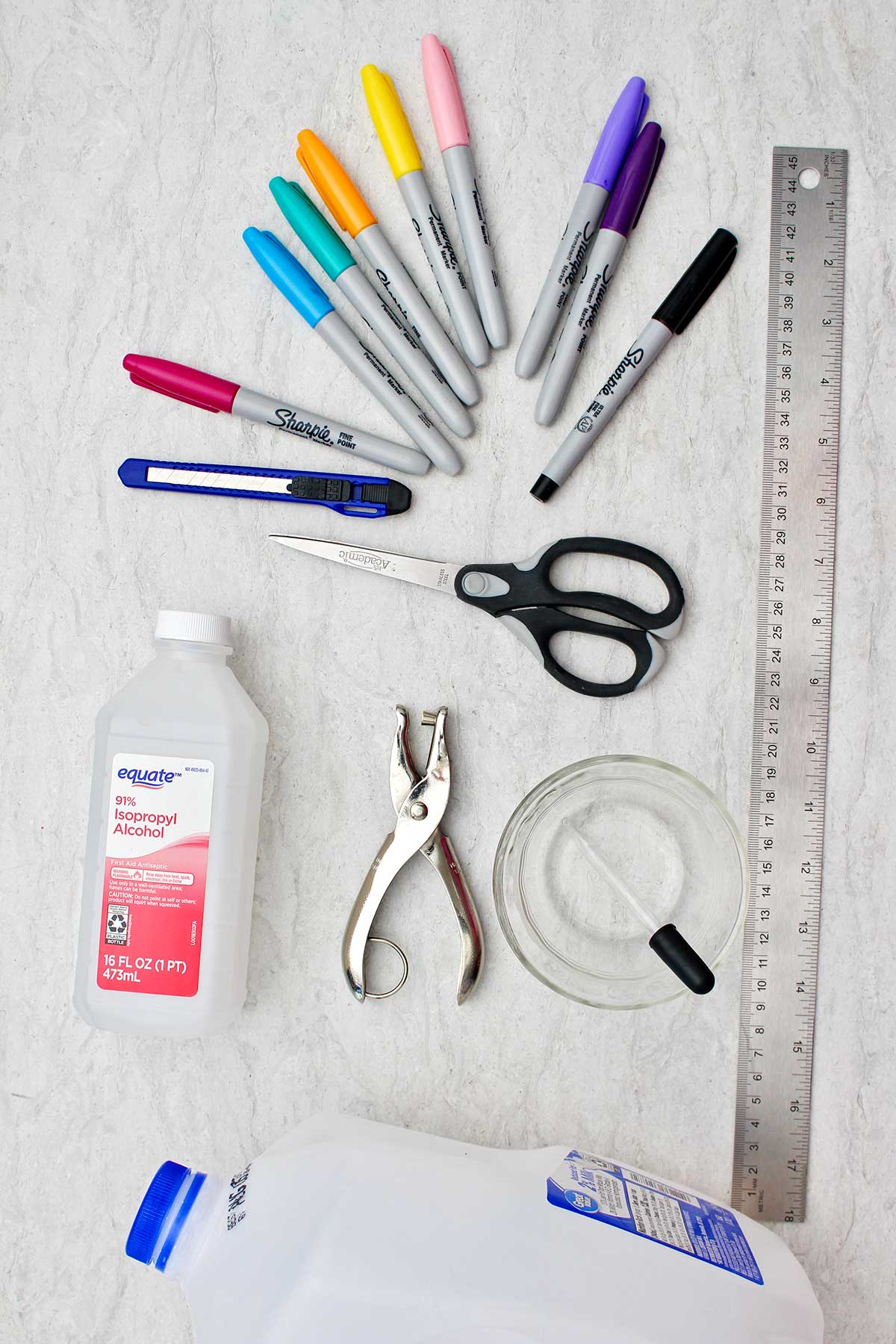 Supplies used to make bookmarks. Colorful sharpies, metal ruler, exact-o knife, scissors, hole punch, alcohol and dropper and plastic milk jug.