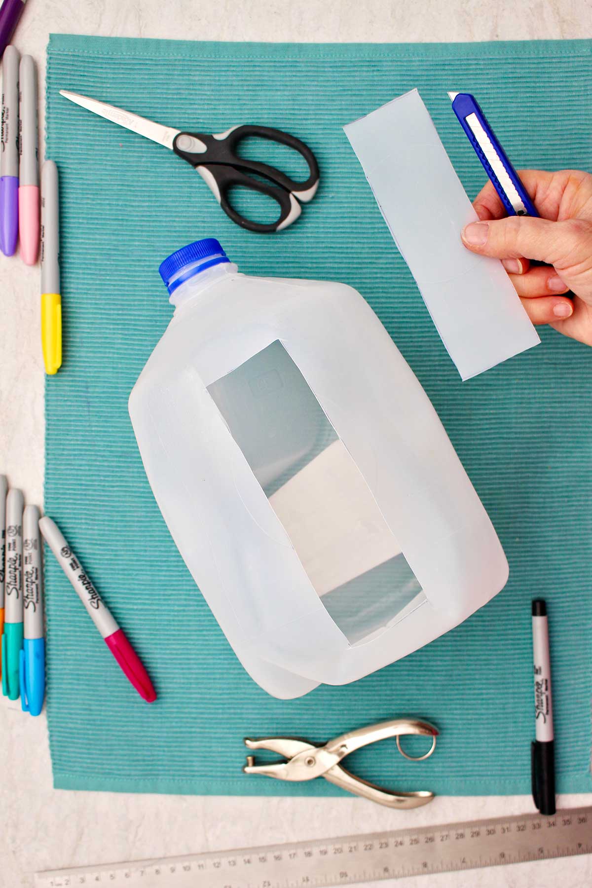 Person holding cut out bookmark from plastic milk jug with sharpies, scissors and other supplies near by.