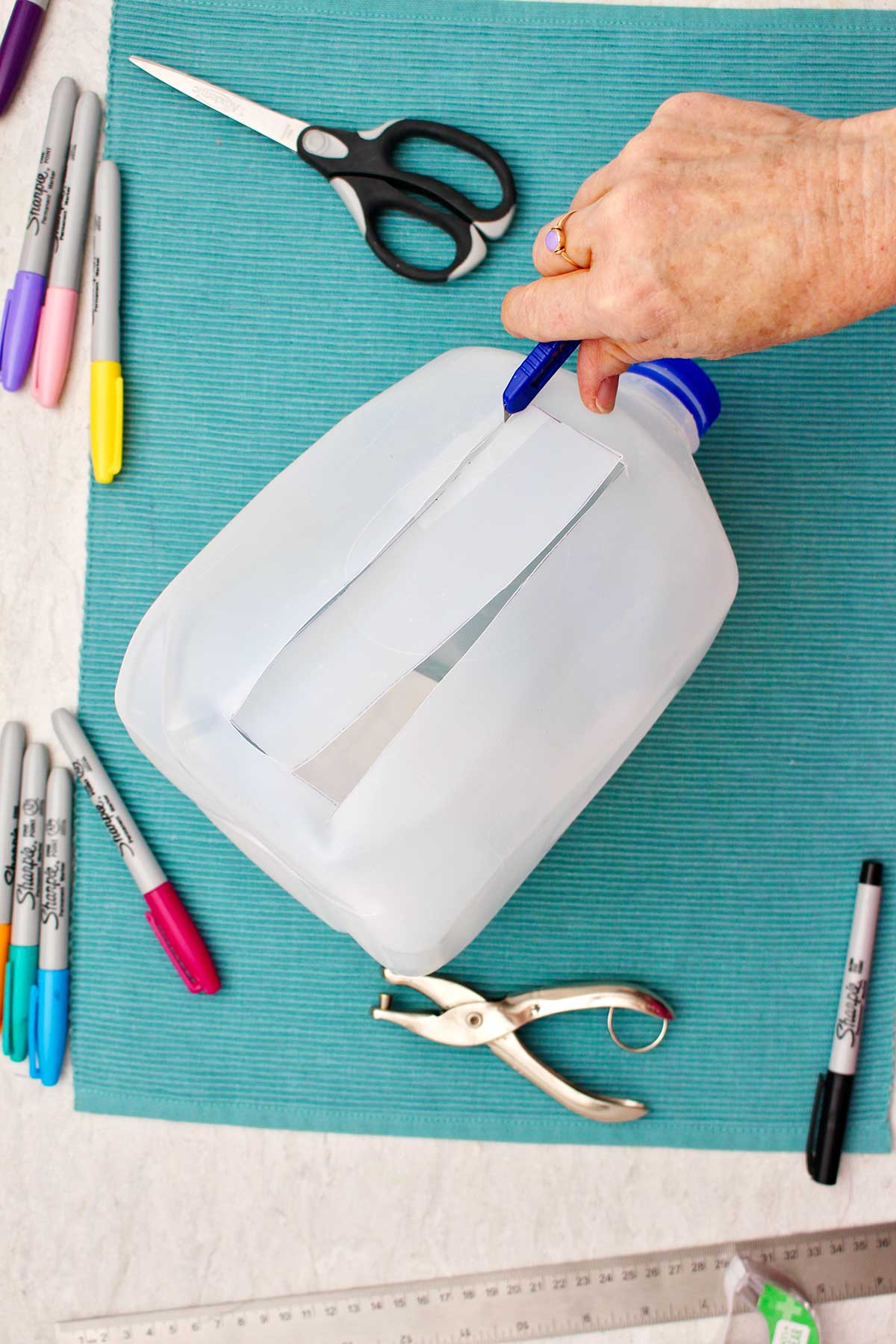 Person cutting bookmark out of plastic milk jug which is resting on a teal placemat.