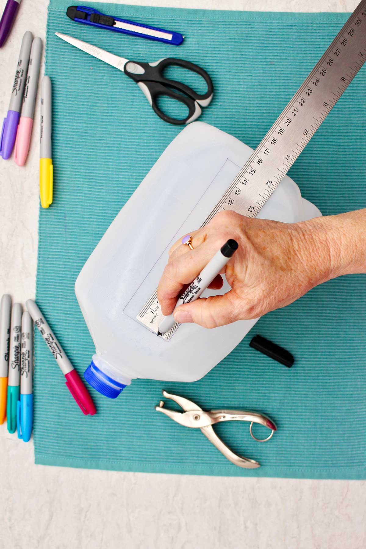 Person marking bookmark shape in plastic milk jug with a sharpie, using a ruler.