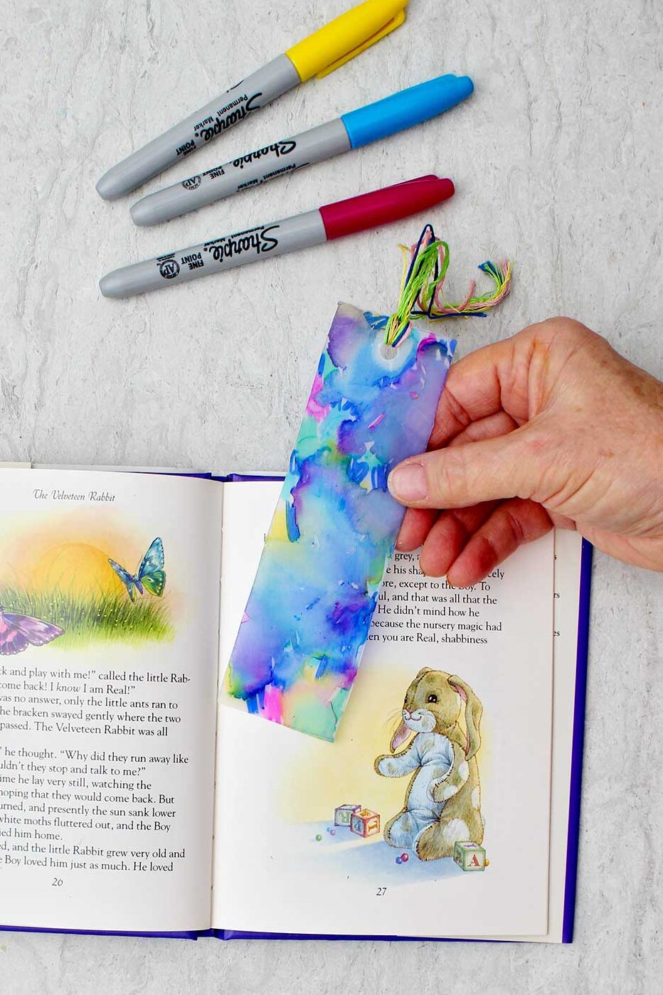 Person holding completed colorful plastic bookmark near an open children's story book with colorful sharpies near by.