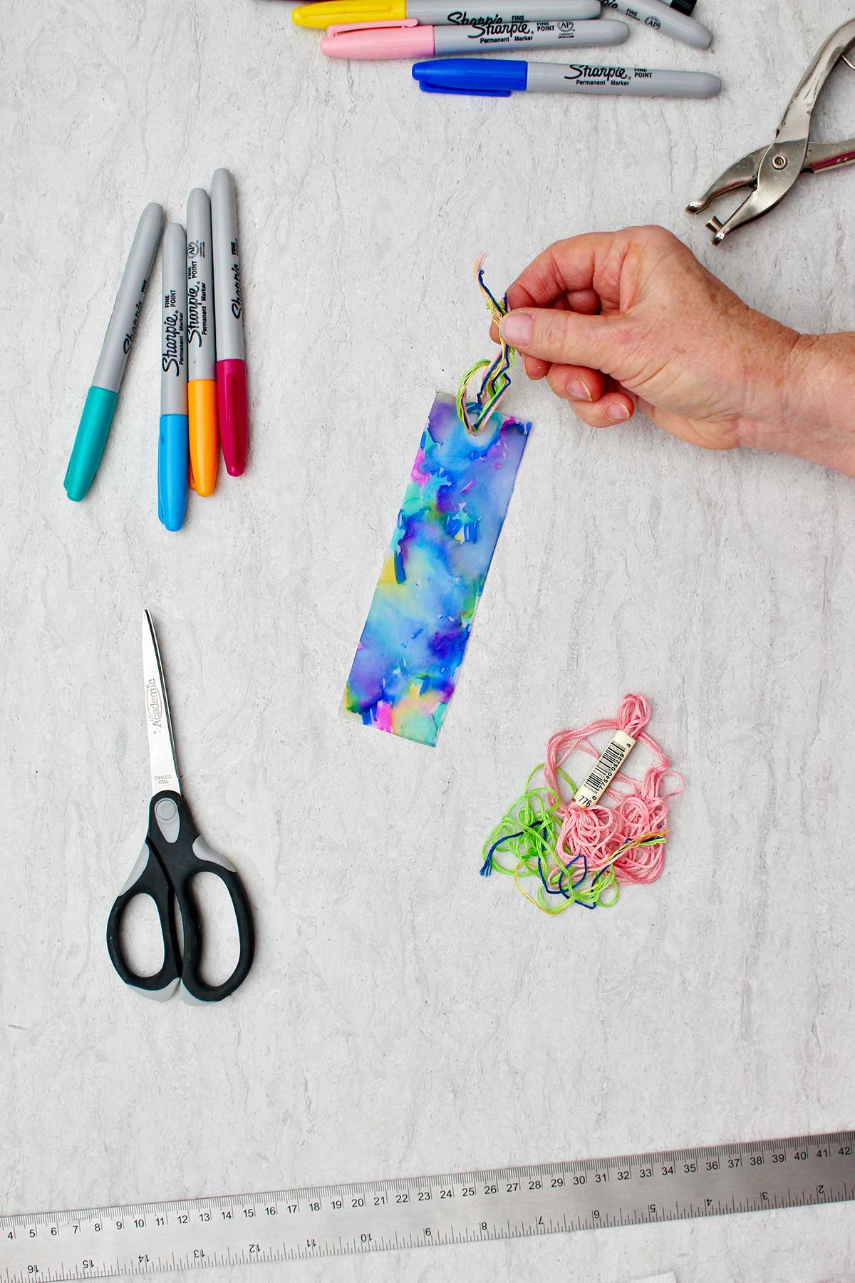 Person securing colorful threads through the hole in the bookmark to make a tassel.