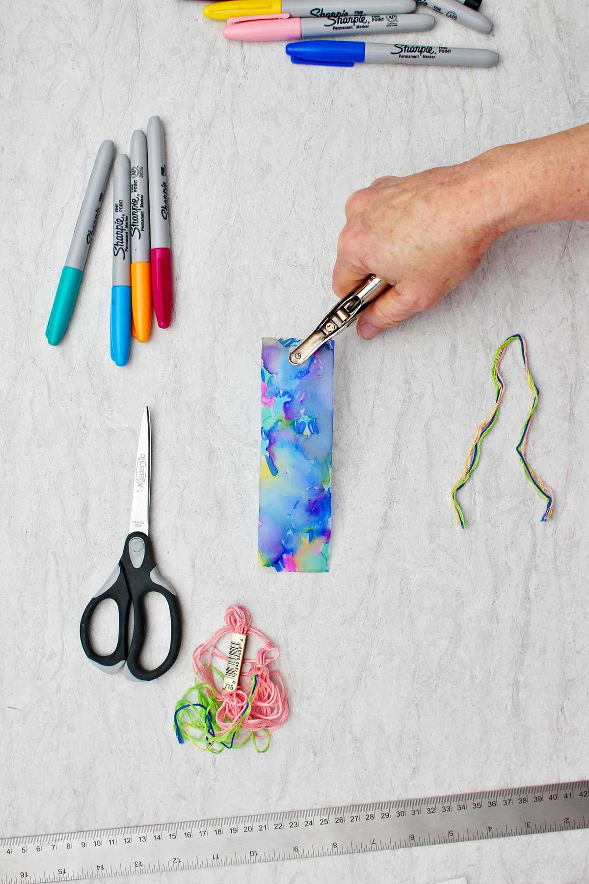 Person punching a hole at the top of a completed colorful plastic bookmark with other supplies near by.