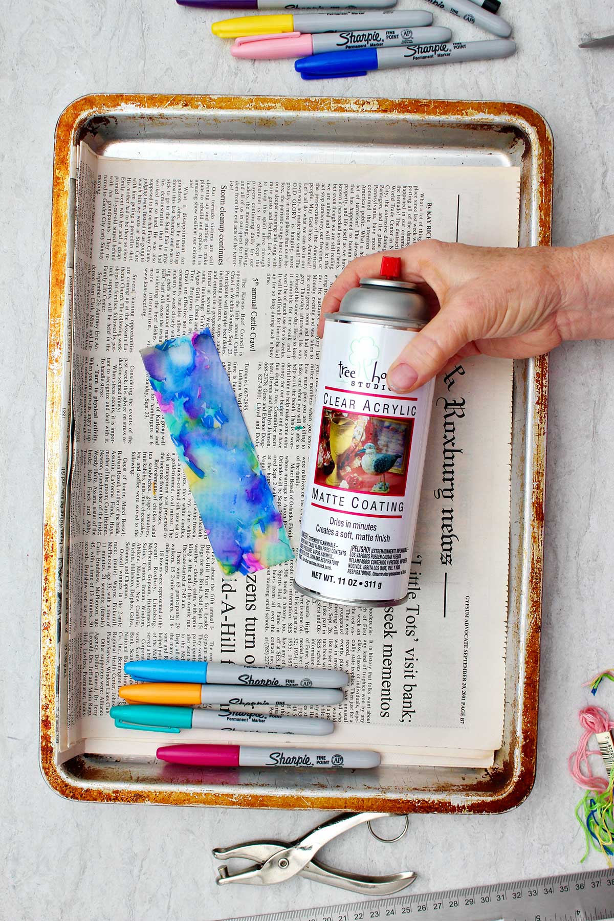 Person holding can of clear acrylic matte coating near partially completed colorful plastic bookmark.