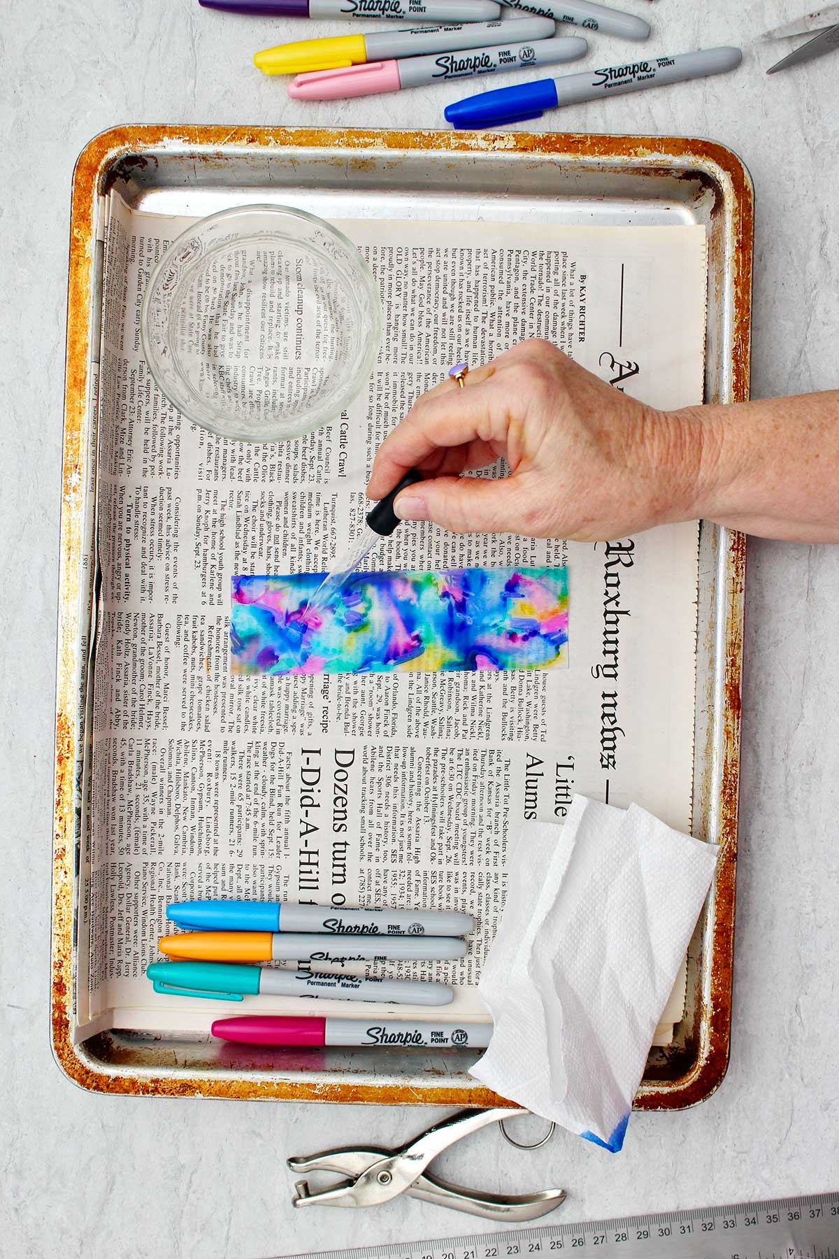 Person using eye dropper to drop alcohol on to colored bookmark resting on a newsprint lined cookie sheet.