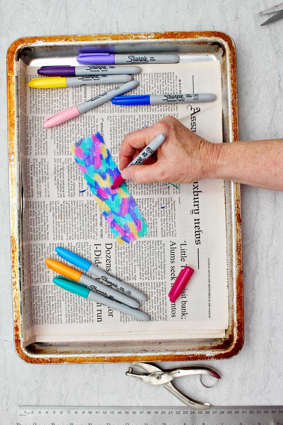 Person coloring plastic bookmark with colorful sharpies on a newsprint lined cookie sheet.