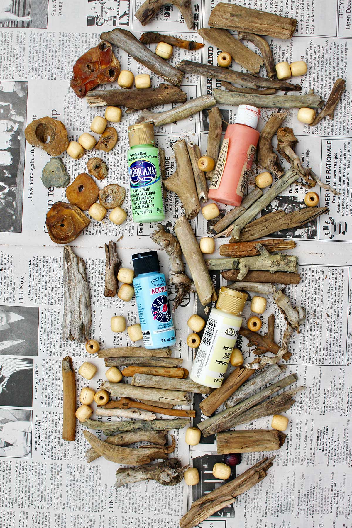 Many pieces of driftwood, acrylic paints and wooden beads on a news print background.