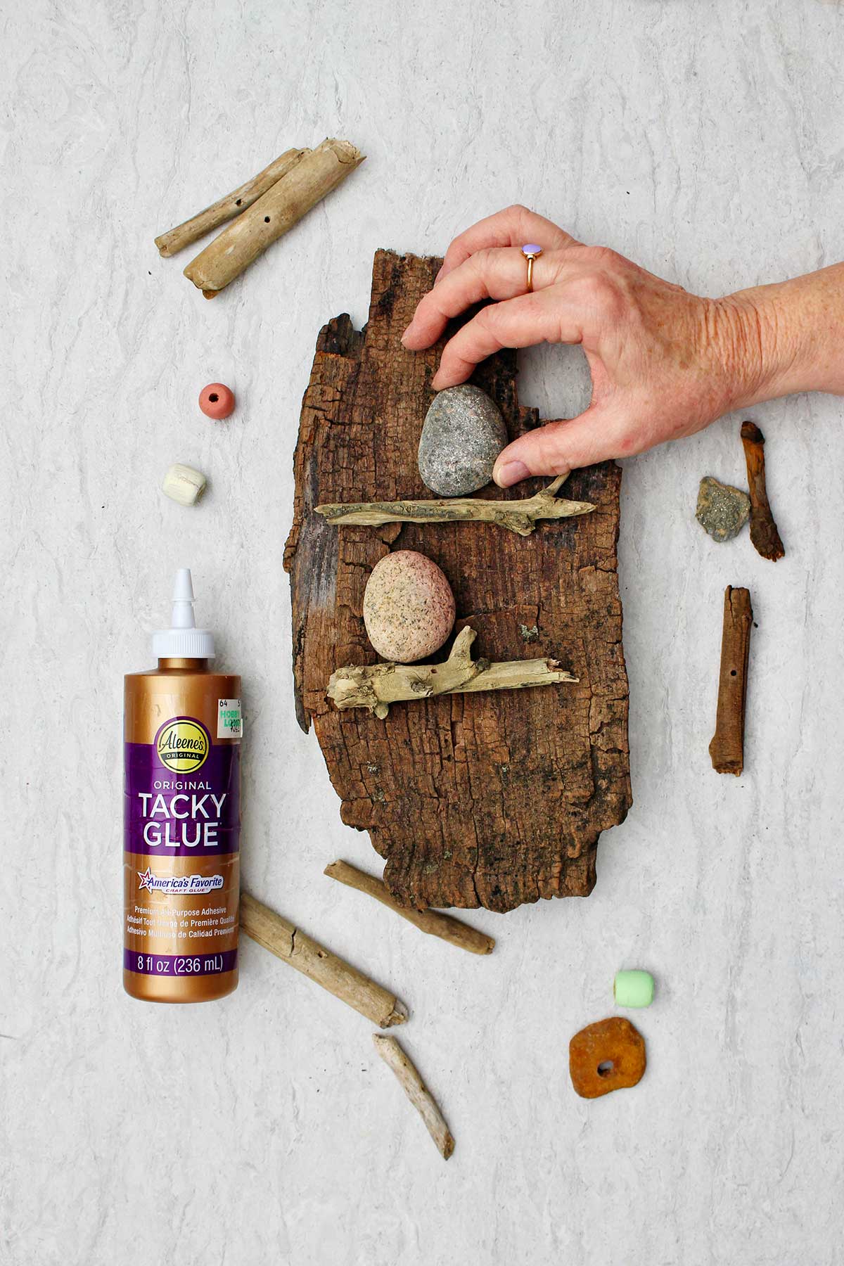 Person placing an egg shaped rock on a driftwood ledge on wooden wall hanging with other pieces of driftwood and tacky glue near by.