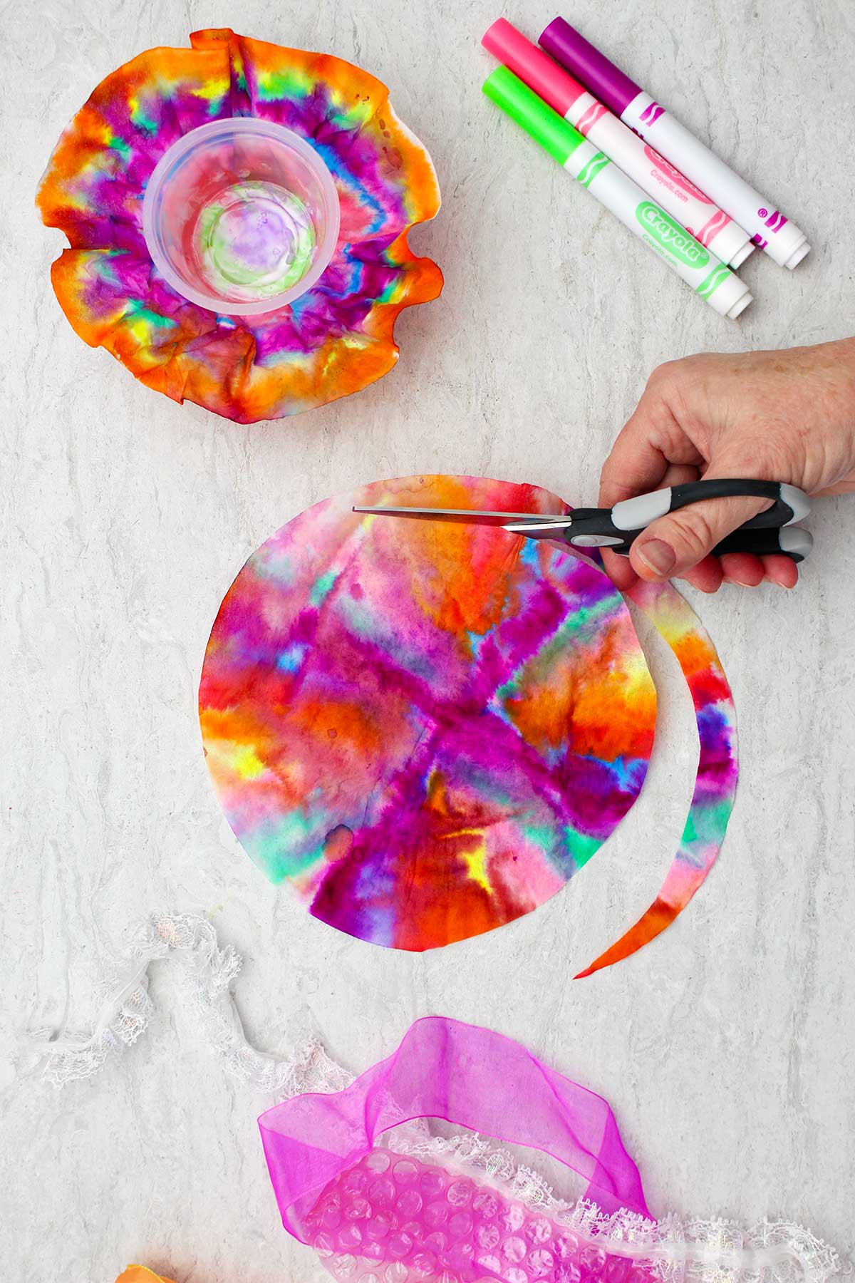 Person cutting a strip around the colored coffee filter with markers and other pieces near by.