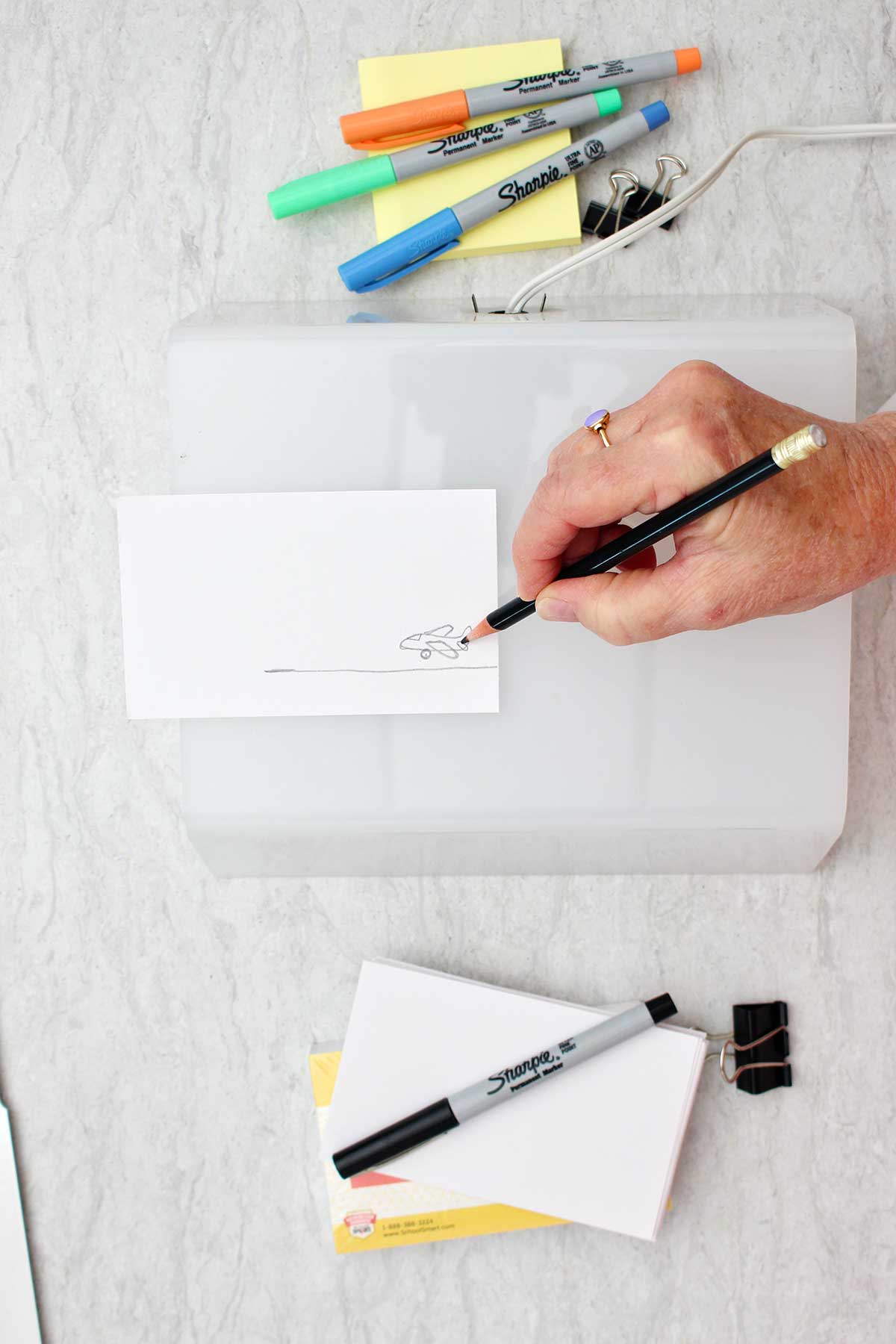 Person sketching a picture of an airplane with pencil on an index card with colorful sharpies and other supplies near by.