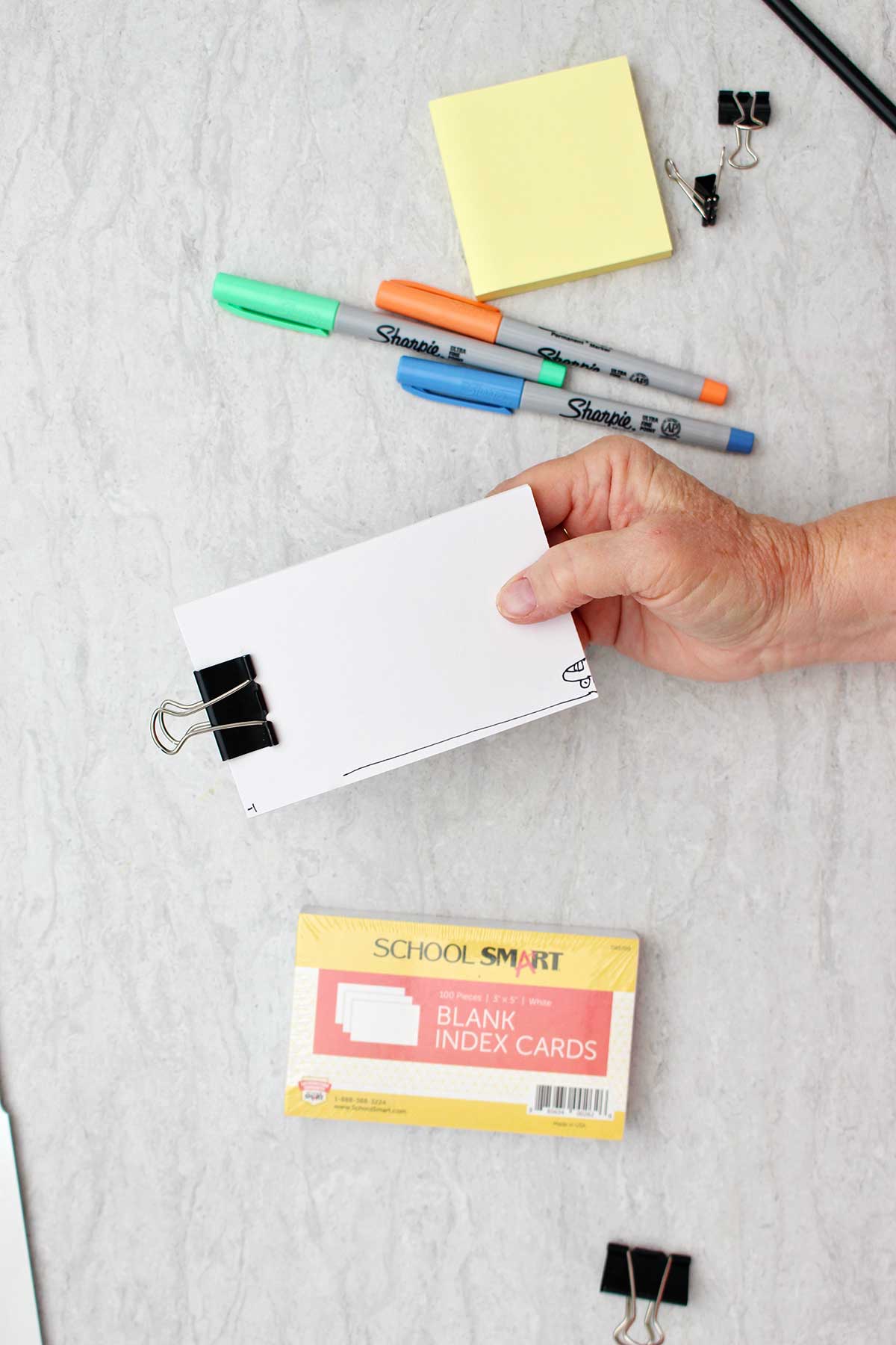 Packet of blank index cards secured with a black binder clip with colorful sharpies and other supplies near by.