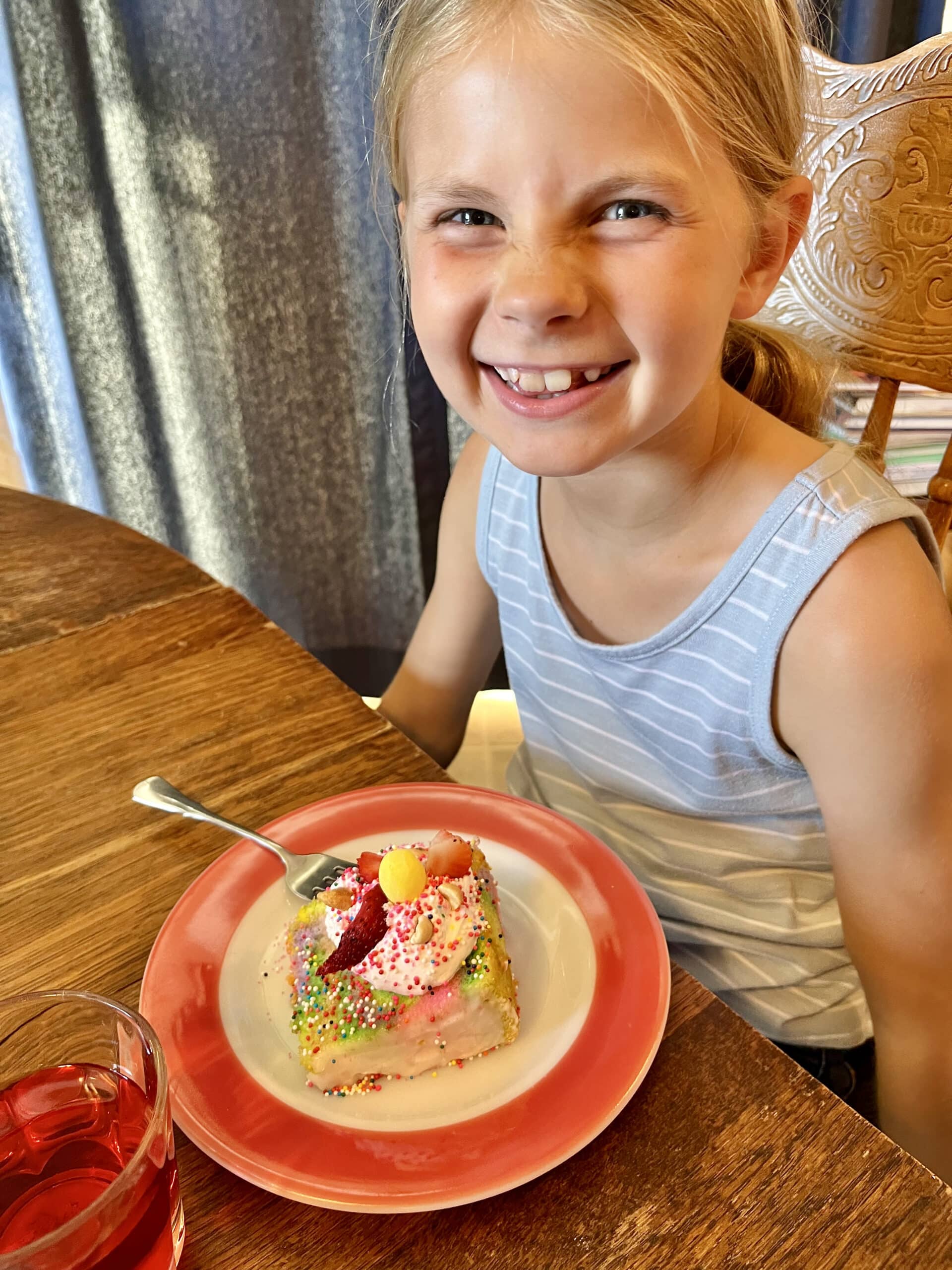 Picture of girl sitting at a table with a piece of angel food cake in front of her. The cake sits on a red and white plate and has a spoonful of whipped cream and sprinkles on top.