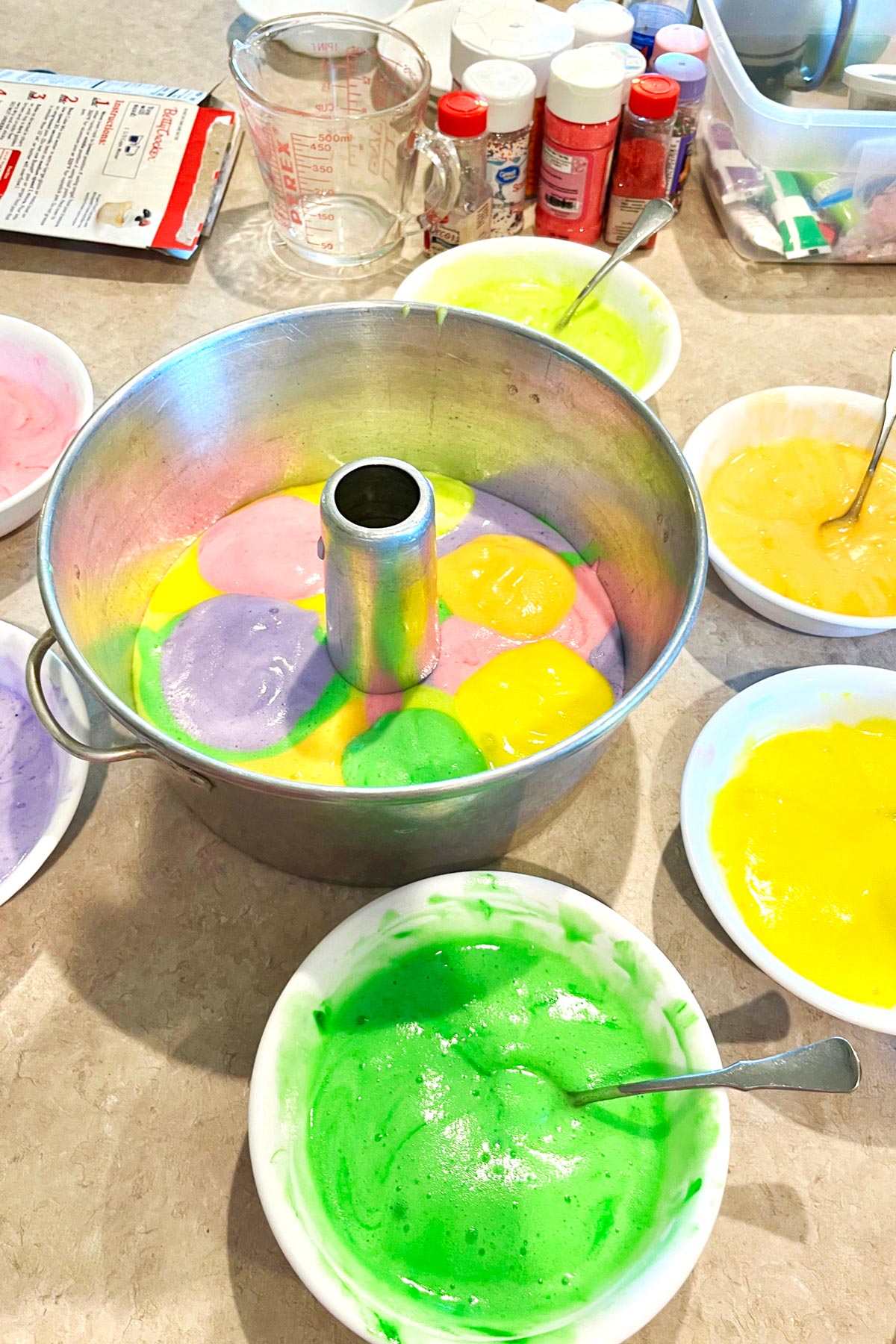 6 bowls of colored cake batter sitting around an angel food cake pan partially full of batter.