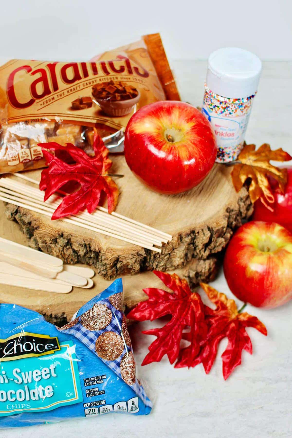 Fall season display of red apples, sticks, semi sweet chocolate chips, caramels, sprinkles and leaves on wood slices.