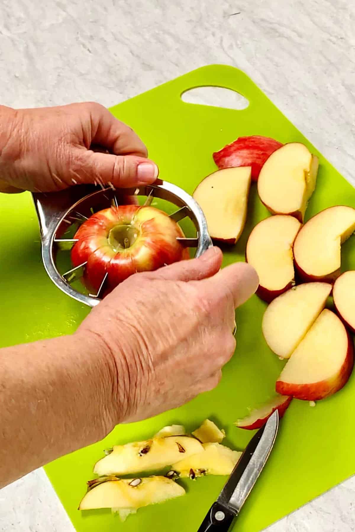 Person pressing apple slicer onto whole apple with other slices nearby on bright green cutting board.