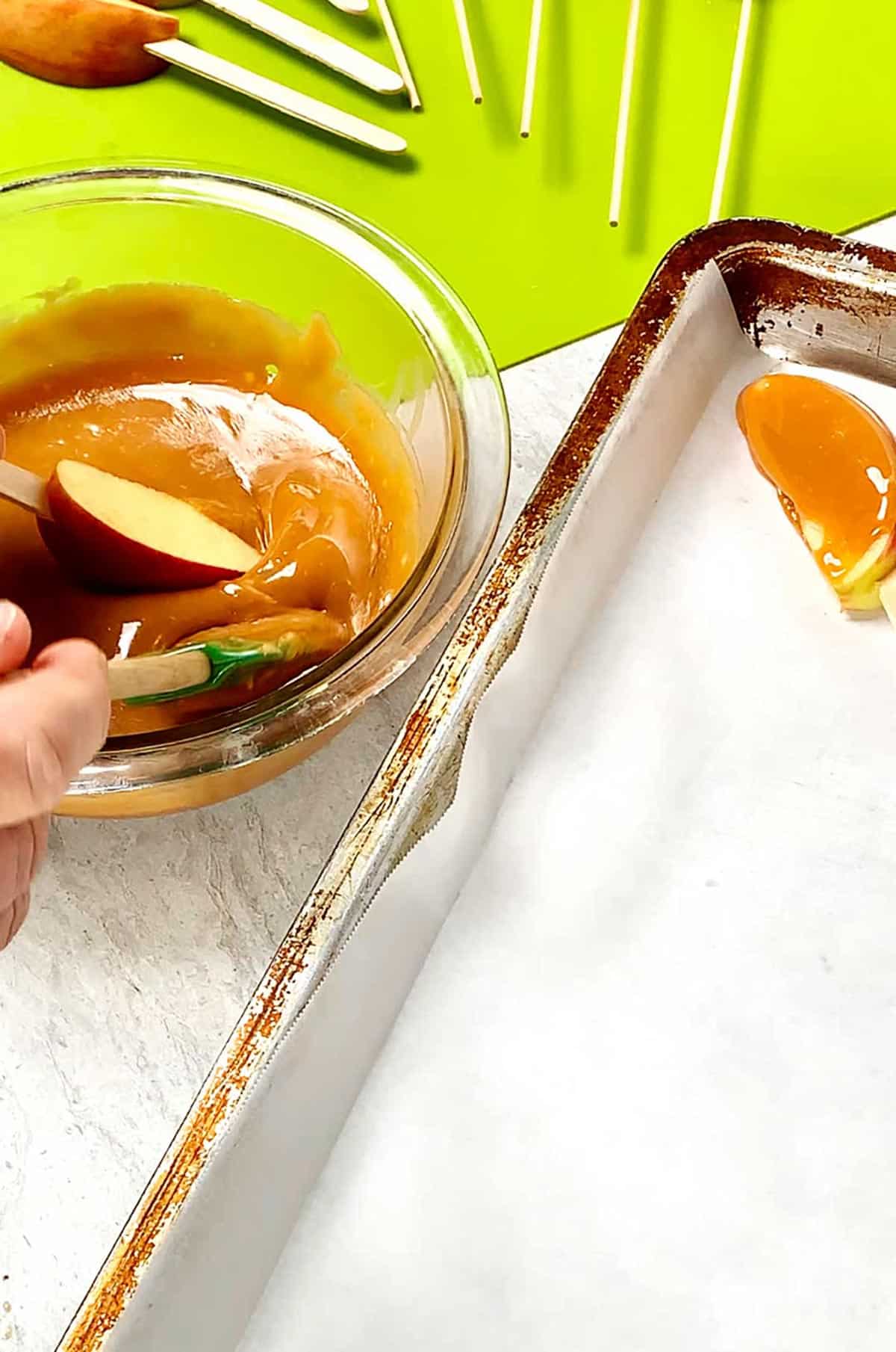 Person dipping apple slices on a stick into warmed caramel in a bowl resting near bright green cutting board and cookie sheet with parchment paper.