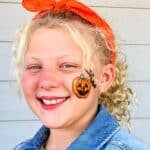 Girl with blonde curly hair smiling at camera with jack-o-lantern face painting on cheek.