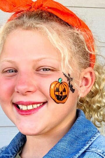 Girl with blonde curly hair smiling at camera with jack-o-lantern face painting on cheek.