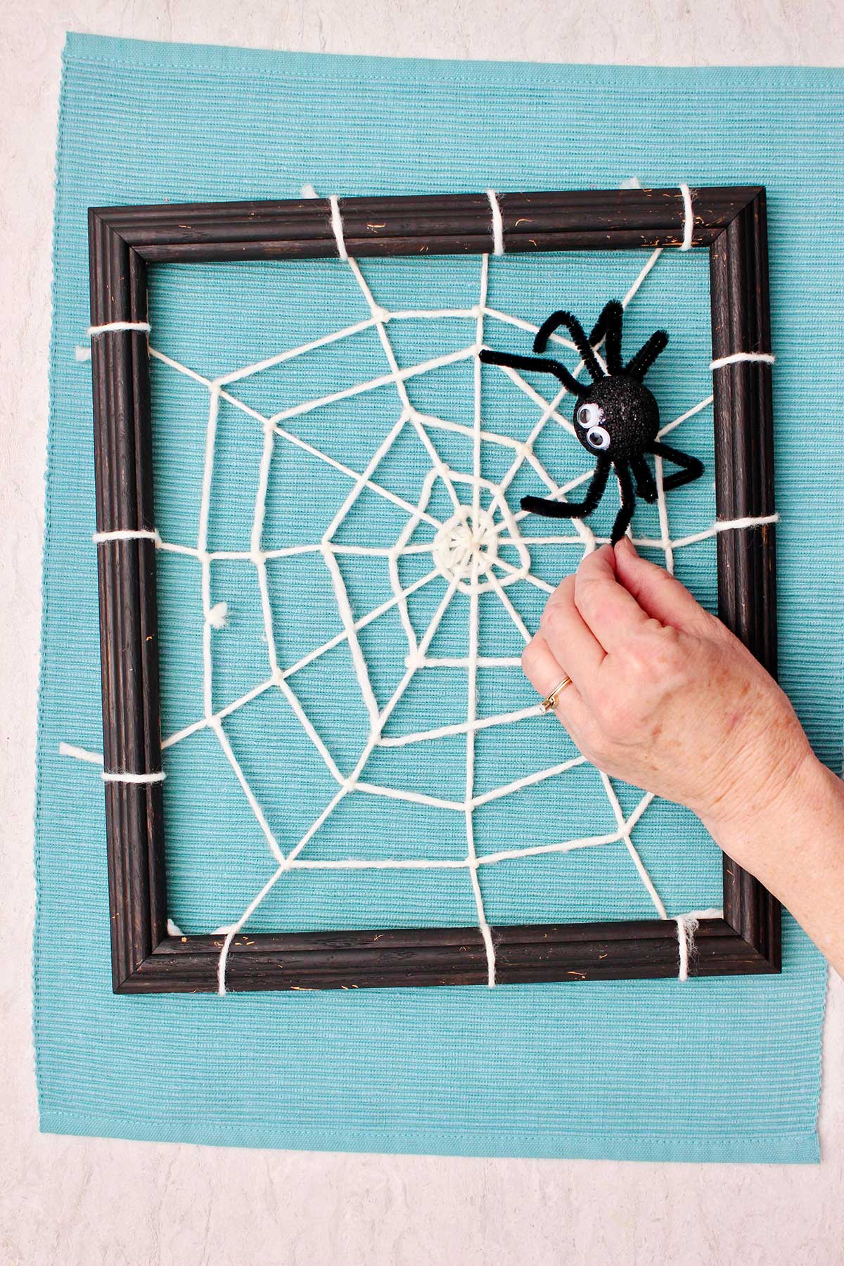 Hand attaching a spider decoration to a DIY spider web made from yarn and attached to a black photo frame resting on an aqua placemat.