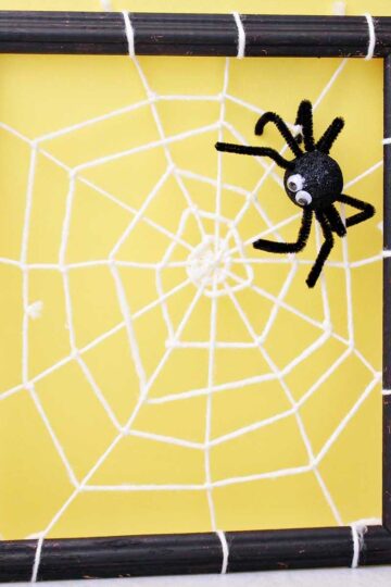 Spider decoration attached to a DIY spider web made from yarn and attached to a black photo frame in front of a yellow back drop.