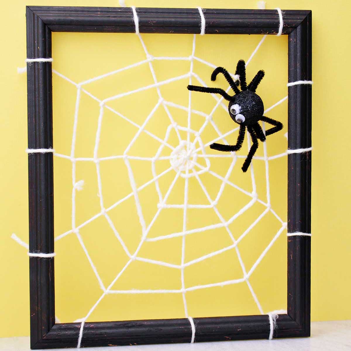 Spider decoration attached to a DIY spider web made from yarn and attached to a black photo frame in front of a yellow back drop.