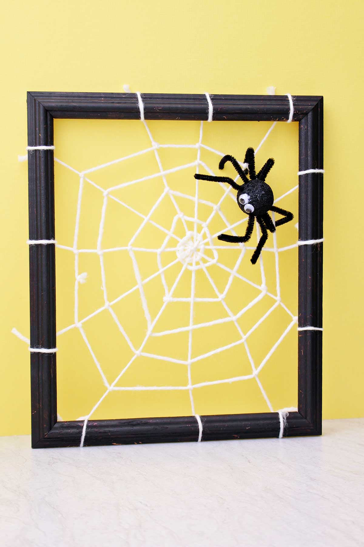 Spider decoration attached to a DIY spider web made from yarn and attached to a black photo frame in front of a yellow back drop.