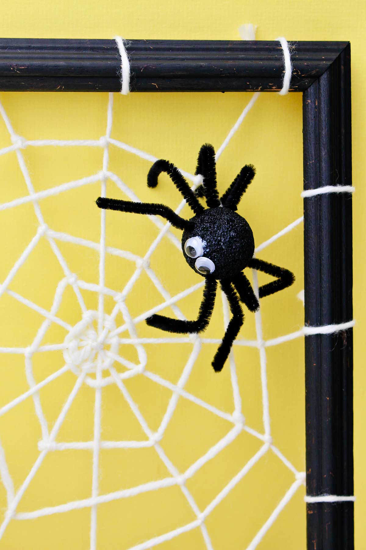 Spider decoration attached to a DIY spider web made from yarn and attached to a black photo frame in front of a yellow back drop.