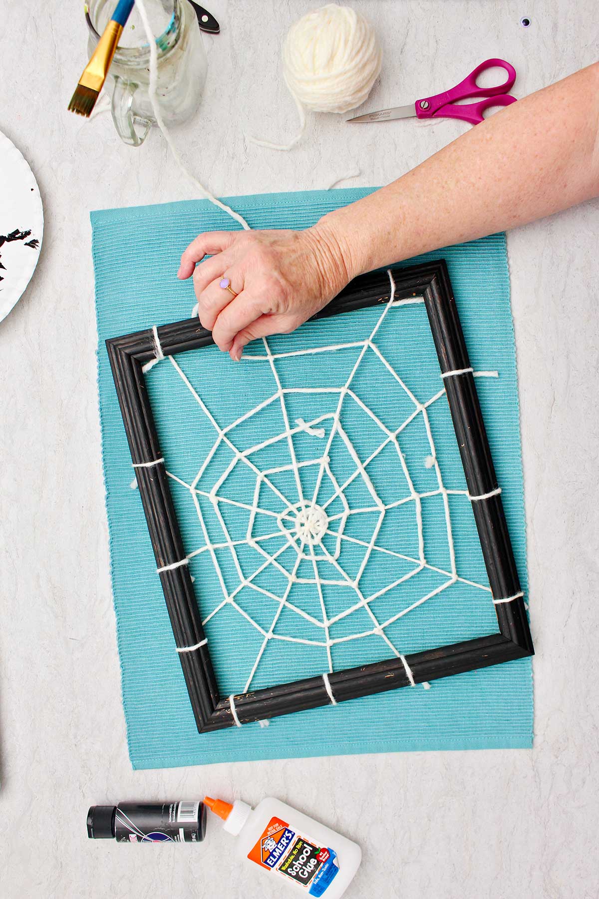 Person making the end of the spider web with white yarn attached to a black picture frame with supplies near by.